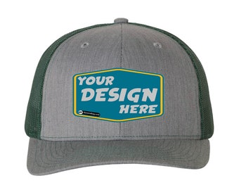 May include: A gray and green trucker hat with a mesh back. The front of the hat has a blue and yellow patch with the text "YOUR DESIGN HERE".