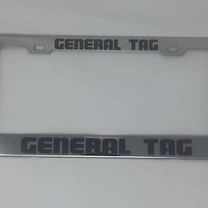 May include: A silver license plate frame with the text "GENERAL TAG" in black lettering on the top and bottom of the frame.