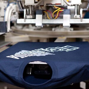 May include: A navy blue long-sleeved t-shirt with a white graphic design featuring the word "NICARAGUA" and a map outline. The shirt is positioned on a screen printing machine, ready for the next step in the printing process.