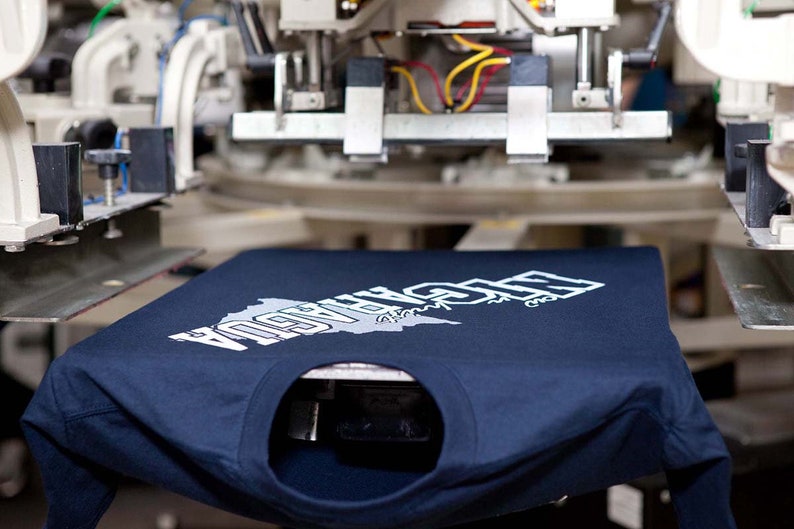 May include: A navy blue long-sleeved t-shirt with a white graphic design featuring the word "NICARAGUA" and a map outline. The shirt is positioned on a screen printing machine, ready for the next step in the printing process.