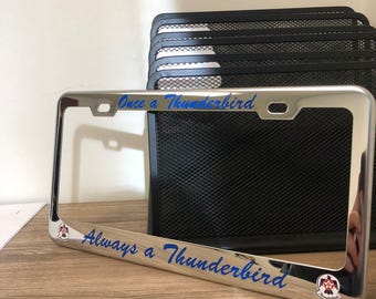 Könnte beinhalten: Ein verchromter Kennzeichenrahmen mit den blauen Worten "Once a Thunderbird" und "Always a Thunderbird". Der Rahmen hat ein kleines Emblem. Ein schwarzer Netz-Organizer und eine Holzoberfläche befinden sich im Hintergrund.