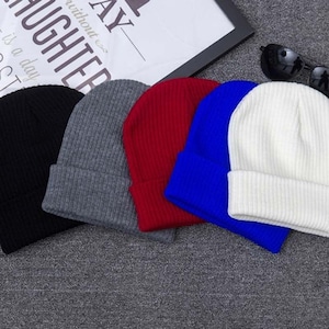 May include: Five knitted beanies in different colors: black, gray, red, blue, and white. The beanies are folded and stacked on top of each other.