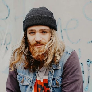 May include: A man with long, wavy, blonde hair wearing a black beanie, a denim jacket, and a grey t-shirt with a red graphic print.