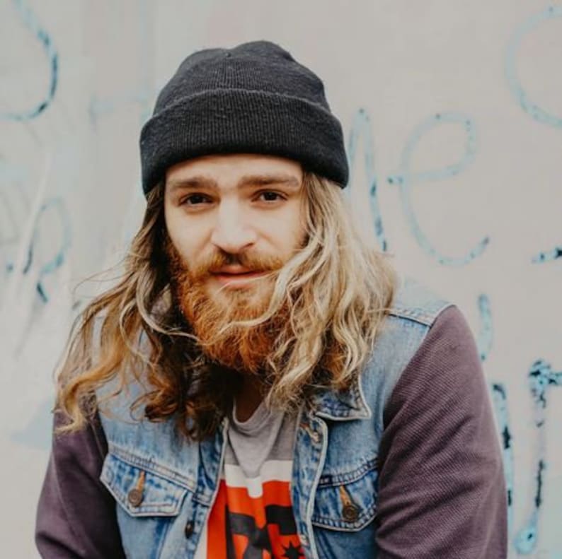 May include: A man with long, wavy, blonde hair wearing a black beanie, a denim jacket, and a grey t-shirt with a red graphic print.