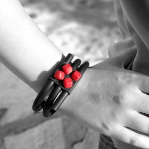 Puede incluir: Un brazalete de cuero negro con cuentas rojas. El brazalete se lleva en la muñeca.
