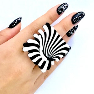 Peut inclure: Une bague noire et blanche avec un motif tourbillonnant et hypnotique. La bague est port&eacute;e sur une main avec un vernis &agrave; ongles noir et blanc.