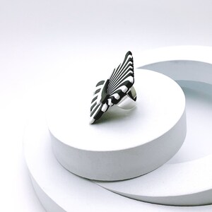 Peut inclure: Une bague &agrave; rayures noires et blanches avec une bande argent&eacute;e. La bague est sur une plateforme blanche.