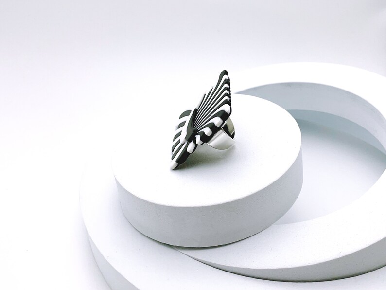 Peut inclure: Une bague &agrave; rayures noires et blanches avec une bande argent&eacute;e. La bague est sur une plateforme blanche.