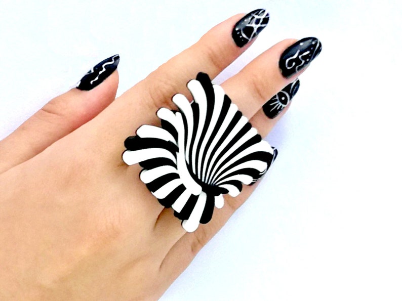 Peut inclure: Une bague noire et blanche avec un motif tourbillonnant et hypnotique. La bague est port&eacute;e sur une main avec un vernis &agrave; ongles noir et blanc.