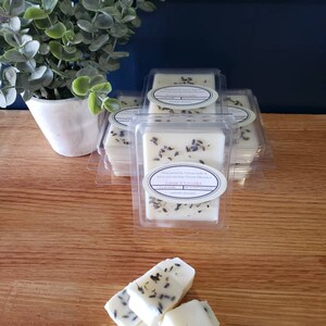 May include: A stack of four white wax melts in clear plastic containers. The wax melts are scented with lemon and lavender and have dried lavender flowers embedded in them. The wax melts are on a wooden surface.