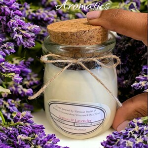 May include: A small glass jar with a cork lid filled with a white candle. The jar is decorated with a brown twine bow and a label that reads "Lemon & Lavender" and "Soy scented candle". The jar is surrounded by purple lavender flowers.