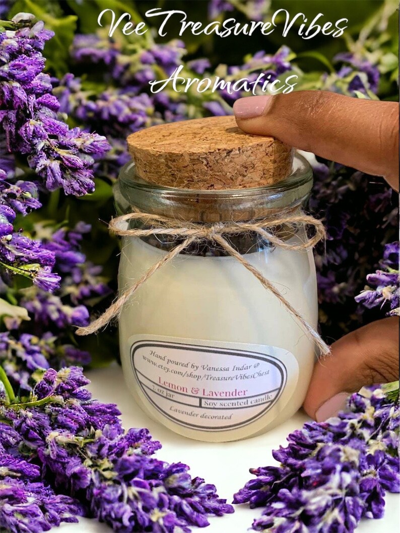May include: A small glass jar with a cork lid filled with a white candle. The jar is decorated with a brown twine bow and a label that reads "Lemon & Lavender" and "Soy scented candle". The jar is surrounded by purple lavender flowers.