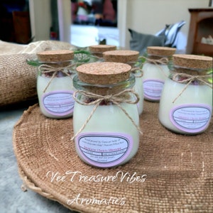 May include: Six small glass jars with cork lids filled with soy wax candles. The jars are arranged on a woven brown surface. The candles are decorated with dried rose petals and twine. The labels on the jars read "Japanese Cherry Blossom" and "Chinese Cherry Blossom" with the text "soy scented candle, sea salt and rose petals infused". The text "Vee Treasure Vibes Aromatics" is printed on the surface.