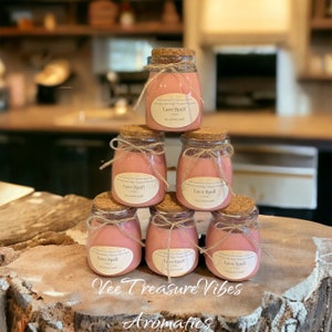 May include: Six pink glass jars with cork lids and twine bows, stacked in a pyramid shape. Each jar has a white label with the text "Love Spell" and "Soy scented candle". The jars are sitting on a wooden surface with the text "Vee Treasure Vibes Aromatics" written below them.