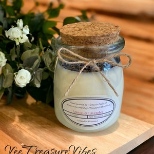 May include: A 7 ounce glass jar candle with a cork lid and a twine bow. The candle is labeled "Lemon & Lavender" and is decorated with dried lavender. The jar is sitting on a wooden surface.
