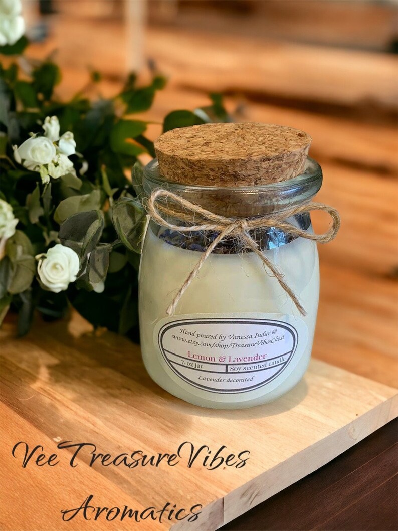 May include: A 7 ounce glass jar candle with a cork lid and a twine bow. The candle is labeled "Lemon & Lavender" and is decorated with dried lavender. The jar is sitting on a wooden surface.