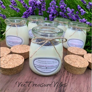 May include: Five glass jars filled with white soy candles decorated with dried lavender. The jars are labeled "Lemon & Lavender" and "Soy Scented Candle". The jars are sitting on a wooden surface with cork lids.