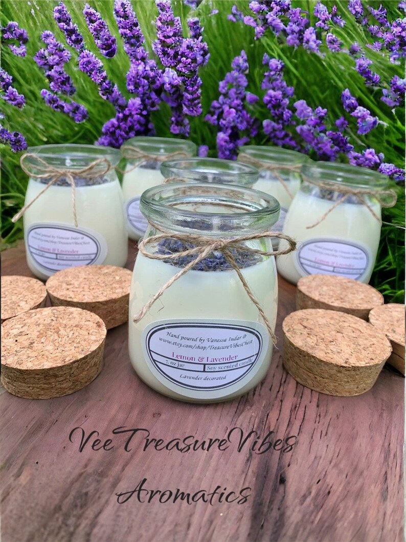 May include: Five glass jars filled with white soy candles decorated with dried lavender. The jars are labeled "Lemon & Lavender" and "Soy Scented Candle". The jars are sitting on a wooden surface with cork lids.
