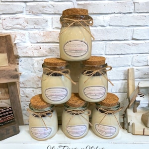 May include: Six glass jars with cork lids and twine tied around the neck, filled with a white candle. The jars are stacked in a pyramid shape on a white surface. The labels on the jars read "Lemon & Lavender" and "Vee Treasure Vibes Aromatics".