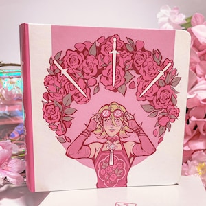 May include: A pink notebook with a floral design featuring a person with pink hair and a rose crown. The notebook has a pink ribbon bookmark and the text "DUYVANNE" on the front cover.