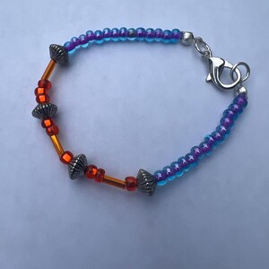 May include: A beaded bracelet featuring a mix of translucent blue and purple seed beads, vibrant orange tube and round beads, and silver-toned metal spacer beads. The bracelet is secured with a silver-toned metal lobster claw clasp.