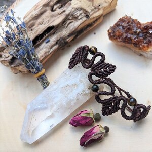 May include: A brown macrame bracelet with tiger's eye beads. The bracelet is on a white surface with a clear quartz crystal, dried lavender, and rose buds.