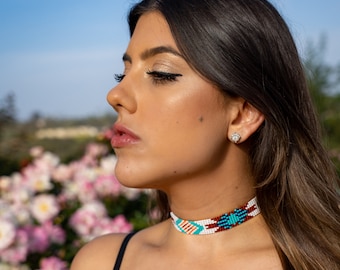 May include: A woman with long dark hair wearing a white, turquoise, and brown beaded choker necklace. The necklace has a geometric pattern.