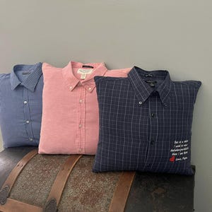 May include: Three decorative pillows made from button-down shirts. One is blue with a checkered pattern, one is pink, and the third is navy blue with a white grid pattern. The navy pillow has embroidered text and a red heart.
