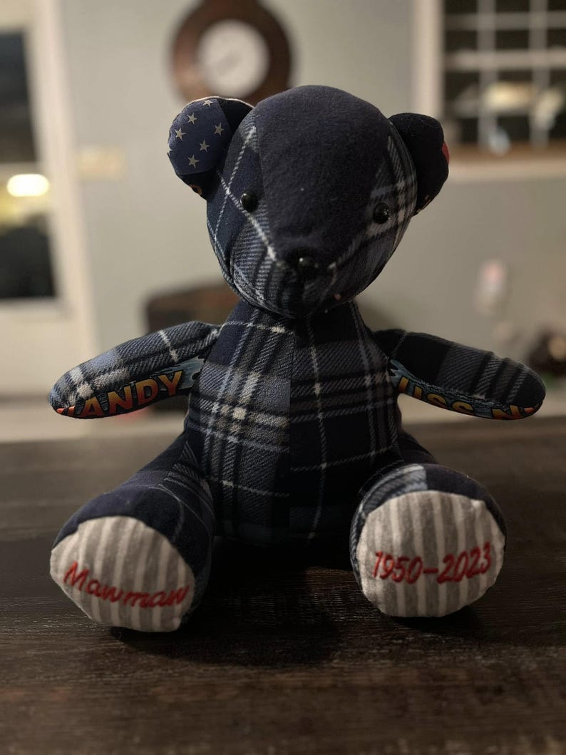 Peut inclure: Un ours en peluche fait &agrave; la main, orn&eacute; d'un motif &agrave; carreaux bleu marine. Le visage de l'ours est noir, avec le mot "ANDY" brod&eacute; sur un bras et les dates "1950-2023" et "Mawmaw" brod&eacute;es sur les pieds. Les oreilles ont un motif d'&eacute;toiles.
