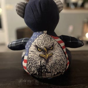 Peut inclure: Un ours en peluche patriotique avec une t&ecirc;te bleu marine et des oreilles ray&eacute;es. La poitrine de l'ours pr&eacute;sente un graphique d&eacute;taill&eacute; d'un aigle avec le drapeau am&eacute;ricain. Les bras de l'ours sont &agrave; carreaux.