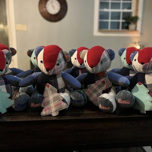 May include: A collection of handmade teddy bears with red, blue, and plaid fabric details. Each bear has a unique design and is accompanied by a small, decorative tree and a round pillow with the word "Maman" or "Papa" embroidered on it.
