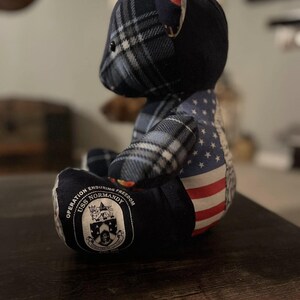 Peut inclure: Un ours en peluche bleu marine avec une t&ecirc;te &agrave; carreaux et un motif de drapeau am&eacute;ricain sur le c&ocirc;t&eacute;. L'ours porte un &eacute;cusson avec l'inscription "Operation Enduring Freedom USS Normandy".