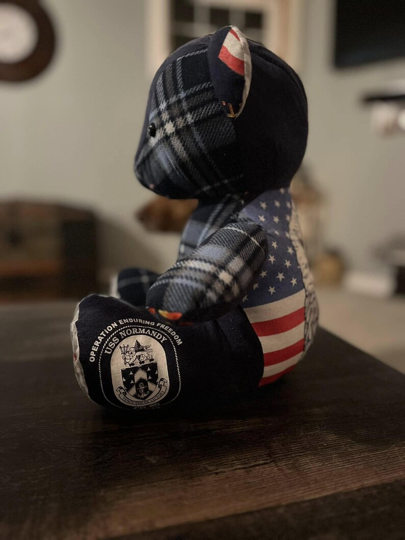 Peut inclure: Un ours en peluche bleu marine avec une t&ecirc;te &agrave; carreaux et un motif de drapeau am&eacute;ricain sur le c&ocirc;t&eacute;. L'ours porte un &eacute;cusson avec l'inscription "Operation Enduring Freedom USS Normandy".