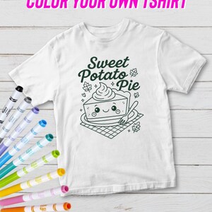 May include: White t-shirt featuring a green graphic of a sweet potato pie slice with a smiling face. The text "Sweet Potato Pie" is above the pie. The words "COLOR YOUR OWN TSHIRT" are at the top. A set of colourful fabric markers are on the left.
