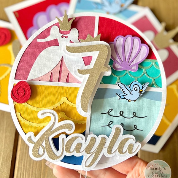 May include: A round cake topper with a gold glitter number 7, a princess, a seashell, a bird, and the name "Kaylie" in gold glitter. The topper is decorated with various colors and textures, including red, yellow, blue, green, and white.