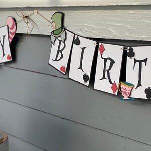 May include: A white and black banner with the words "Happy Birthday" decorated with playing cards and Alice in Wonderland characters. A pink frosted cake with a topper featuring Alice in Wonderland characters sits in front of the banner.