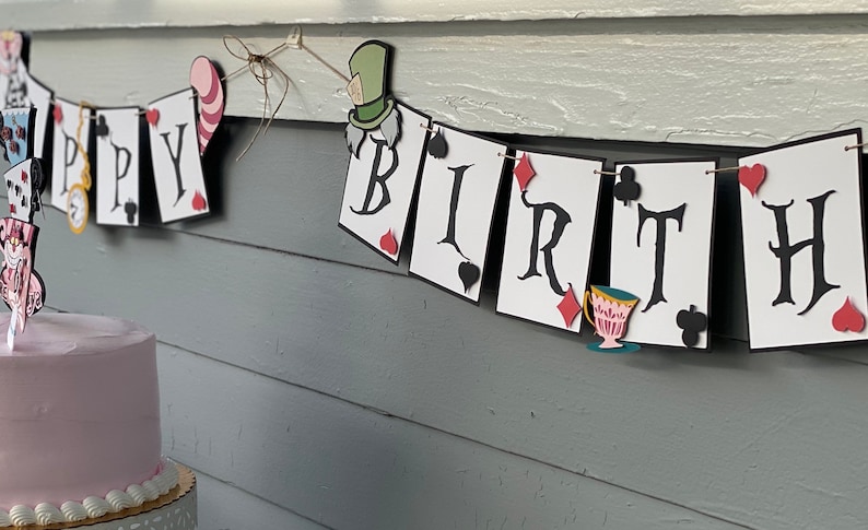 May include: A white and black banner with the words "Happy Birthday" decorated with playing cards and Alice in Wonderland characters. A pink frosted cake with a topper featuring Alice in Wonderland characters sits in front of the banner.