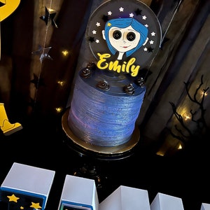 May include: A blue and black cake with a white frosting and a black and white character topper. The topper has the name "Emily" written in yellow letters. The cake is on a black cake stand with a gold trim. The cake is in front of a black background with white stars.
