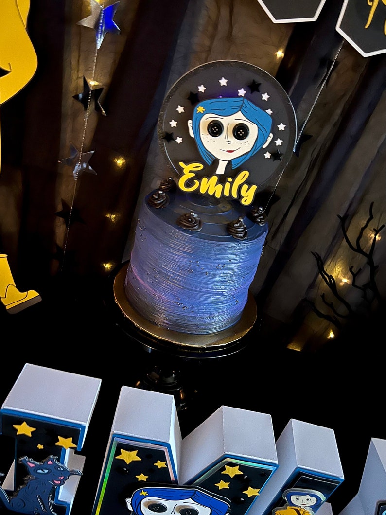 May include: A blue and black cake with a white frosting and a black and white character topper. The topper has the name "Emily" written in yellow letters. The cake is on a black cake stand with a gold trim. The cake is in front of a black background with white stars.