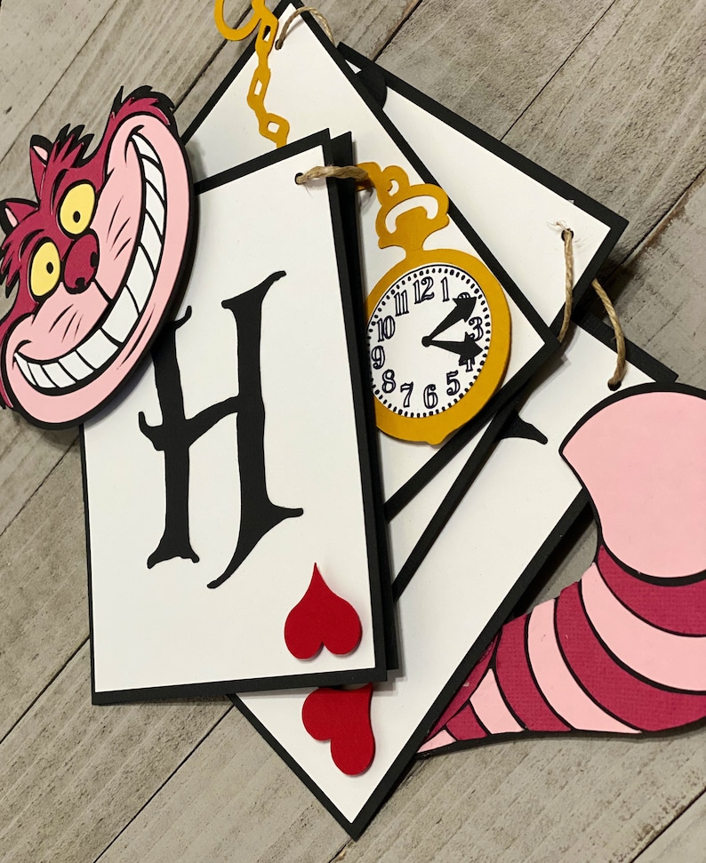May include: Alice in Wonderland themed party banner with a Cheshire Cat, playing cards, hearts, and a pocket watch. The banner says "H" for Happy.