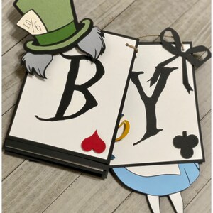 May include: A white banner with the words "BY" in black, decorated with a Mad Hatter character, playing cards, and a blue and white Alice in Wonderland character.