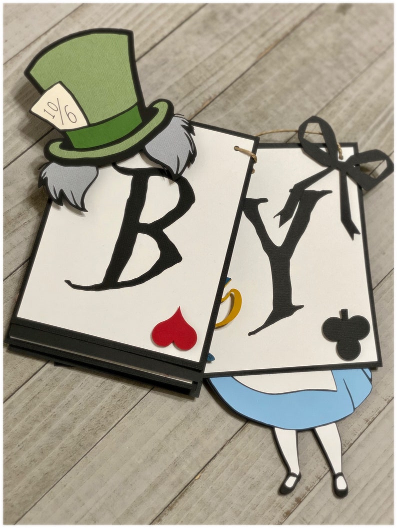 May include: A white banner with the words "BY" in black, decorated with a Mad Hatter character, playing cards, and a blue and white Alice in Wonderland character.