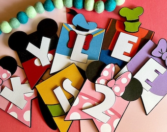 May include: A colourful banner featuring Mickey Mouse, Minnie Mouse, Donald Duck and Goofy characters. The banner spells out "Kylie is 2" in white letters on a variety of coloured backgrounds.