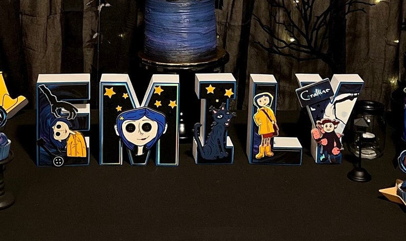 May include: White foam letters spelling "EMILY" decorated with characters from the movie "Coraline". The letters have a black background with blue and yellow accents and stars. The letter "E" features a hand reaching out from a black hole, the letter "M" features Coraline, the letter "I" features a black cat, the letter "L" features Coraline in a yellow raincoat, and the letter "Y" features a character from the movie "Coraline" holding a sign that says "Coraline".