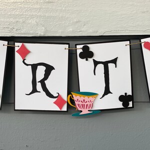 May include: A white banner with black letters spelling out "PARTY" with a teacup and saucer, red and black playing card symbols, and a string running through the top.