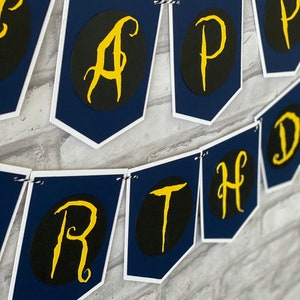 May include: A navy blue and white banner with the word "Happy Birthday" written in gold script letters. Each letter is on a separate flag.