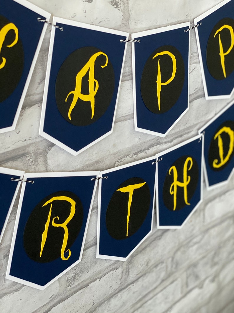 May include: A navy blue and white banner with the word "Happy Birthday" written in gold script letters. Each letter is on a separate flag.