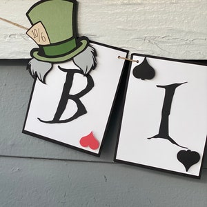 May include: A banner with the letters "BI" in black on white cards with black borders. The letter "B" has a red heart in the corner and the letter "I" has a black spade in the corner. A Mad Hatter character with a green hat and white hair is attached to the left side of the banner.