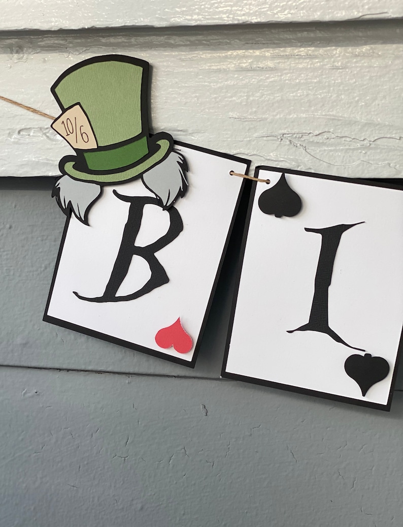 May include: A banner with the letters "BI" in black on white cards with black borders. The letter "B" has a red heart in the corner and the letter "I" has a black spade in the corner. A Mad Hatter character with a green hat and white hair is attached to the left side of the banner.