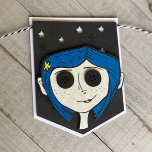May include: A black and white banner with a cutout of a girl with blue hair and large black button eyes. The banner has a black background with white stars cut out.